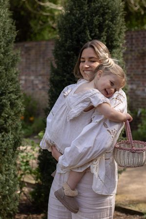 A smiling woman in white dress holds a laughing toddler girl with a wicker basket in a lush garden in Brighton