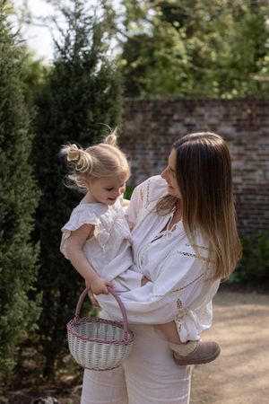 A mother holds her toddler daughter, both in white outfits, as the child holds a wicker basket outdoors during a family session in Brighton