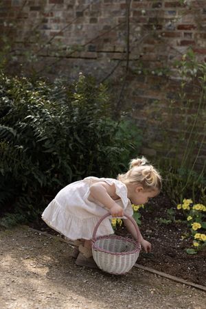 Young girl in white dress crouching down with wicker basket, picking flowers near brick wall during a family portrait session in Brighton
