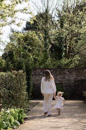 A mother and young daughter in white outfits walk hand-in-hand along a garden path surrounded by lush greenery in Brighton
