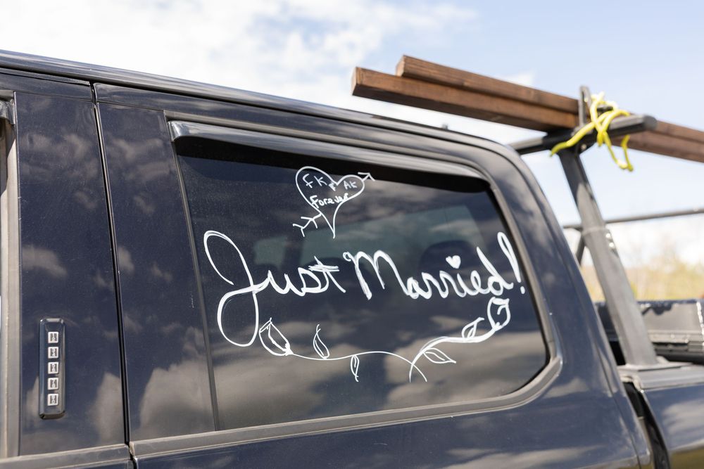 Dark truck window with white Just Married decal and cartoon birds, parked outdoors with lumber in truck bed.
