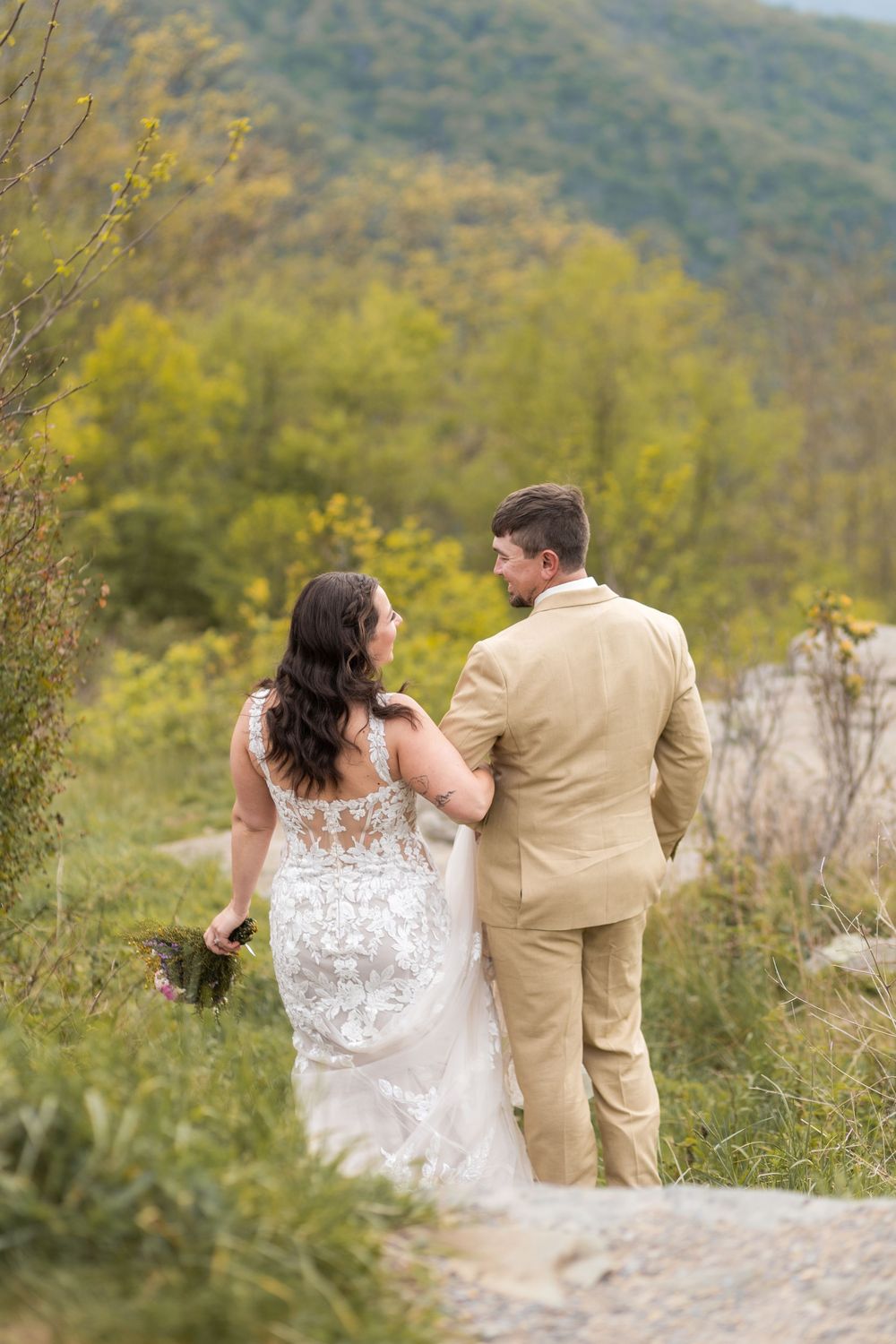 A bride in lace gown and groom in tan suit walk hand-in-hand on a scenic mountain path surrounded by lush greenery.