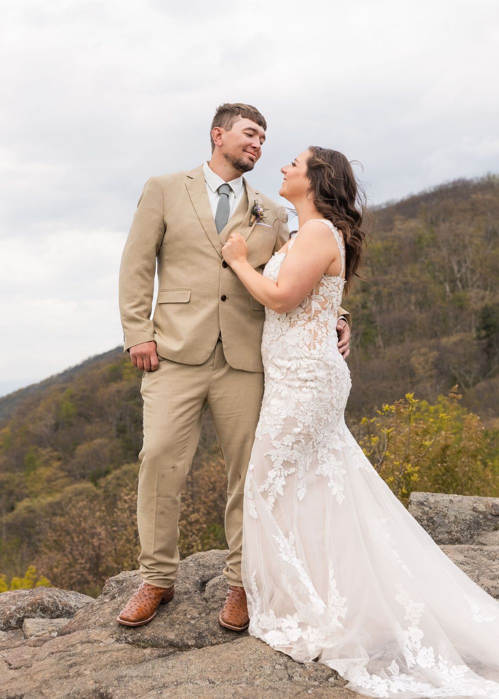 Bride in lace gown and groom in tan suit share a romantic moment on a rocky overlook with mountain views.