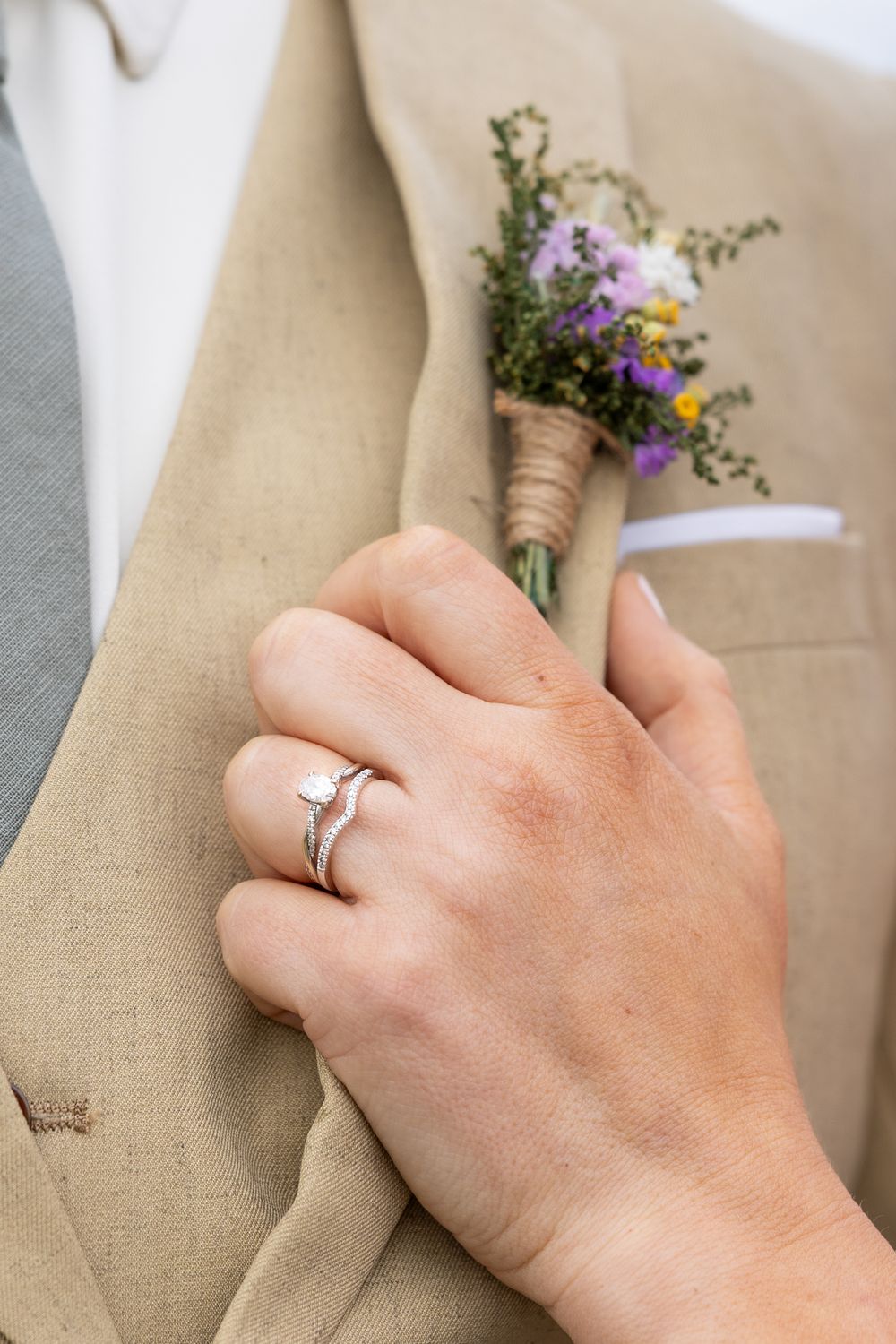 Bride's hand with diamond engagement ring and wedding band resting on groom's tan suit with wildflower boutonniere.