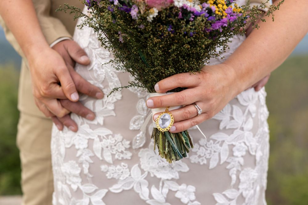 Close-up of bride holding wildflower bouquet wearing lace wedding dress and floral ring with groom.