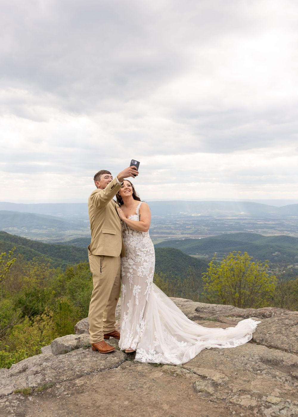 Bride and groom taking a selfie on a mountain overlook, with sweeping valley views, wearing wedding attire on their special day.