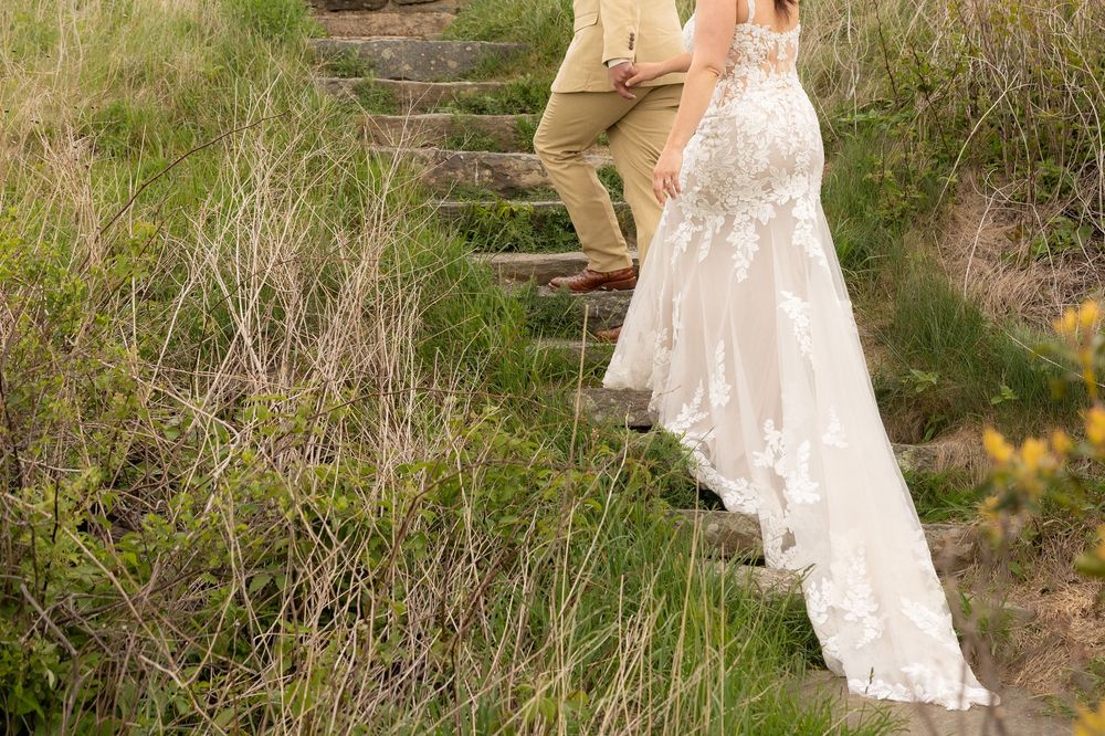 Bride in lace wedding gown and groom in tan suit walking up stone steps surrounded by wild grass.