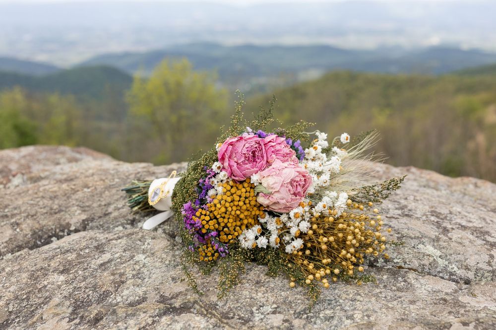A colorful wildflower bouquet with pink roses and yellow blooms resting on a rocky ledge with a scenic mountain valley backdrop.