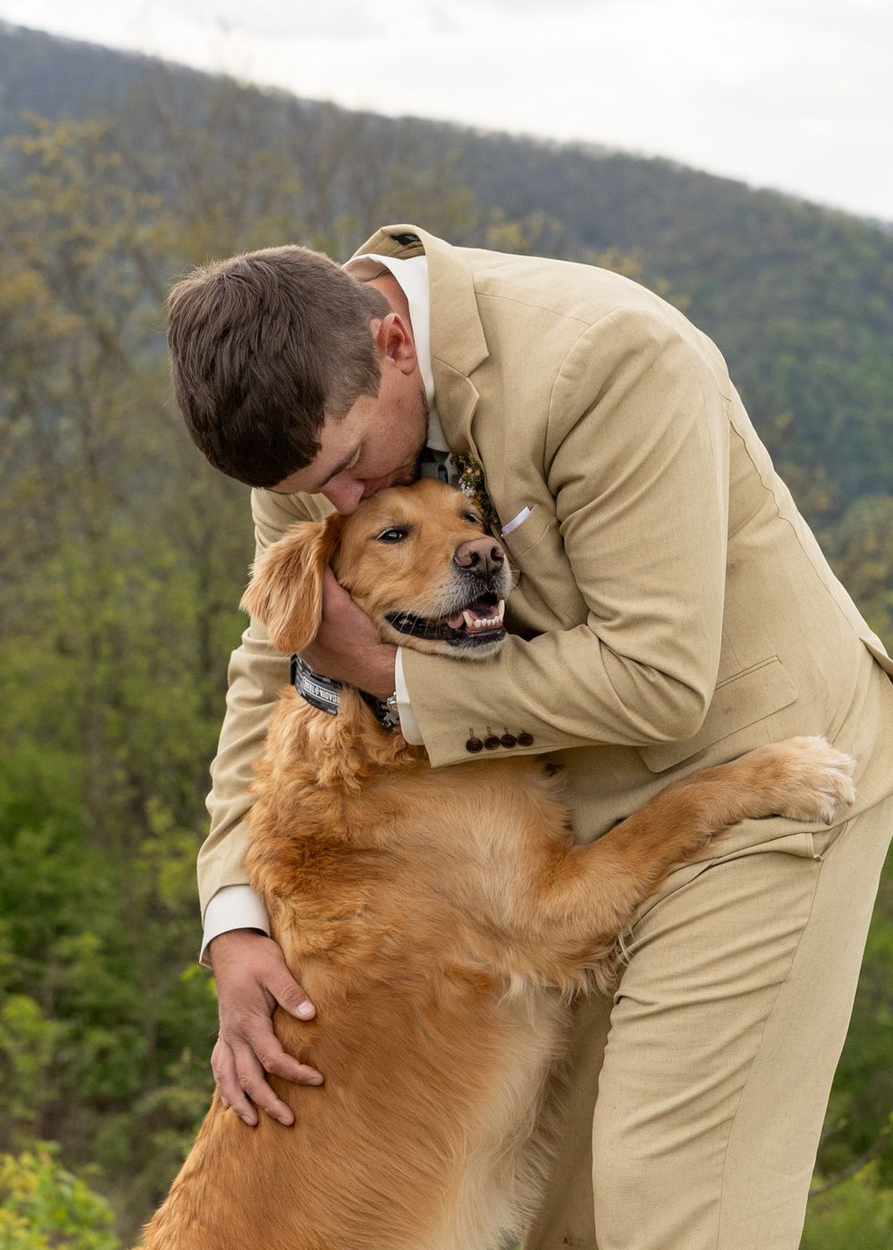 A groom in a tan suit hugs his golden retriever dog outdoors with green mountains in the background.