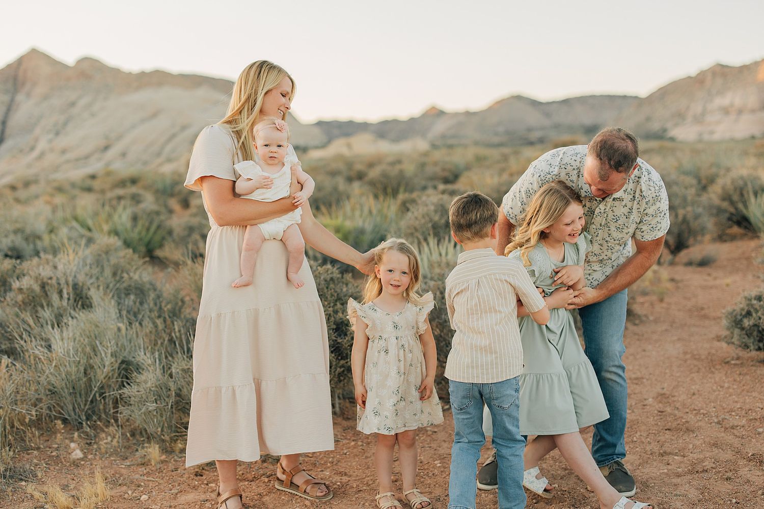 Family of six posing outdoors in desert landscape at golden hour, parents with four children in soft neutral tones.