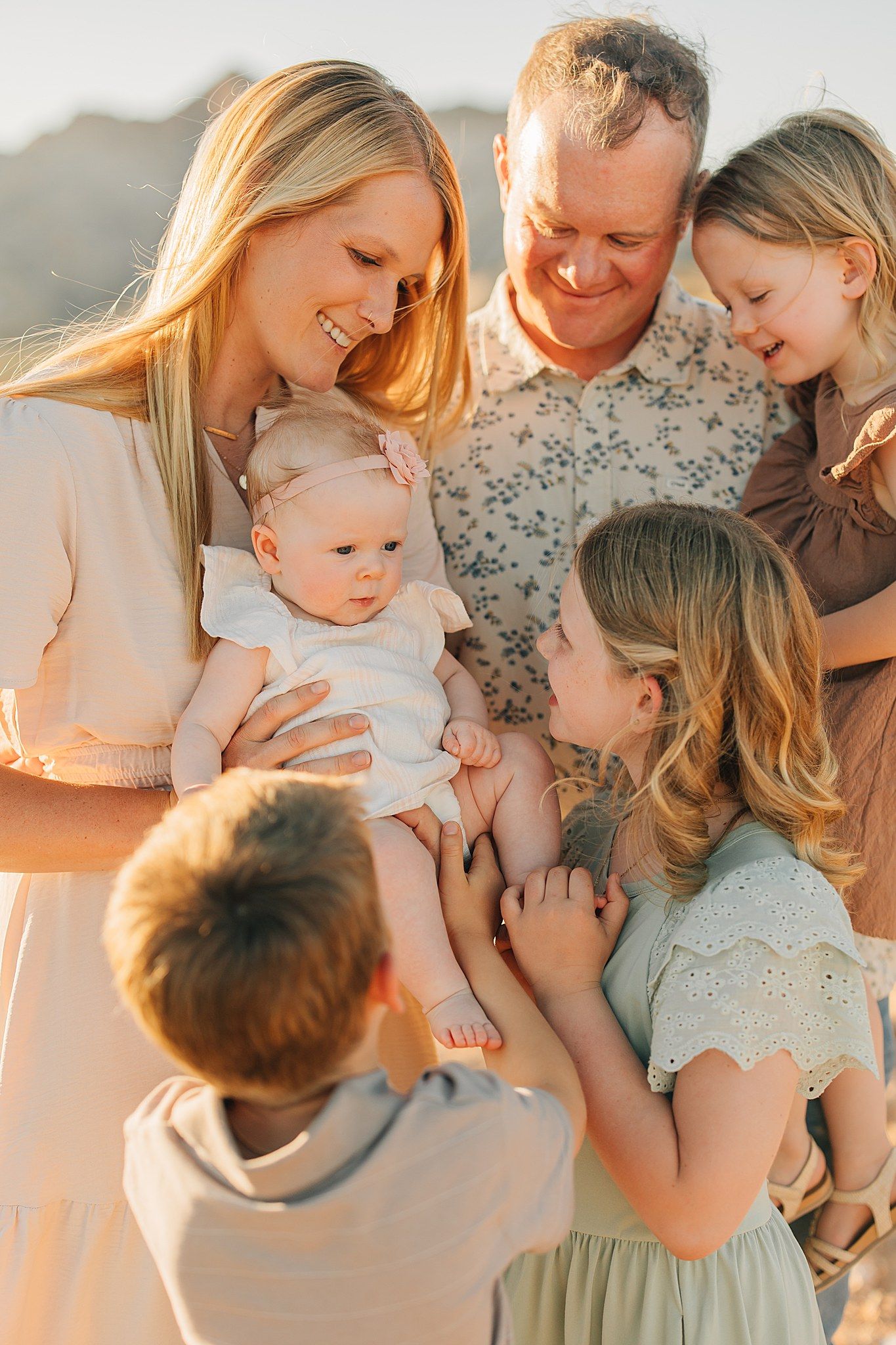 Family of six gathered closely outdoors at golden hour, parents smiling at newborn baby surrounded by three young children.