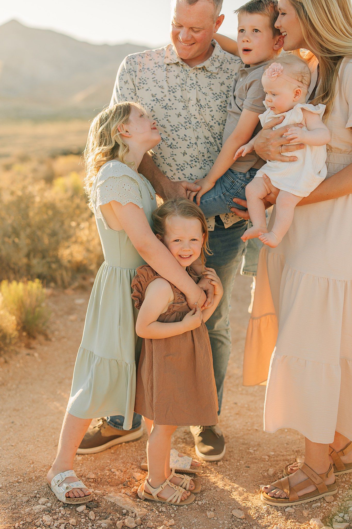 A joyful family of five poses together in a golden desert landscape at sunset, with children laughing and embracing.
