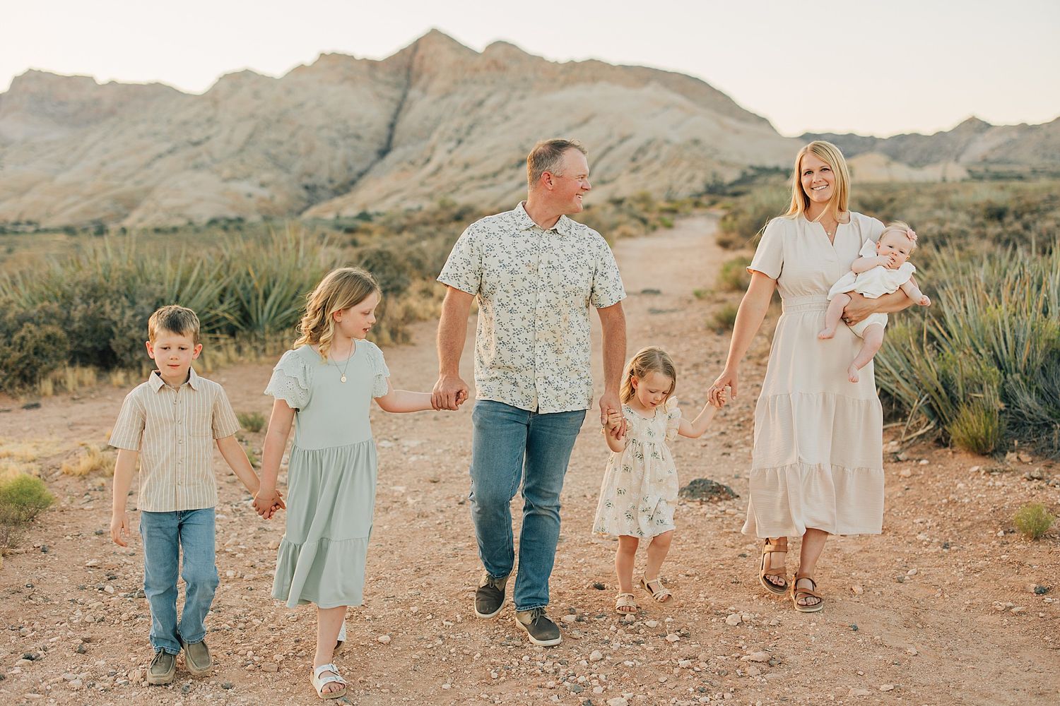 Family of six walks hand-in-hand on a desert path with mountain backdrop during golden hour.