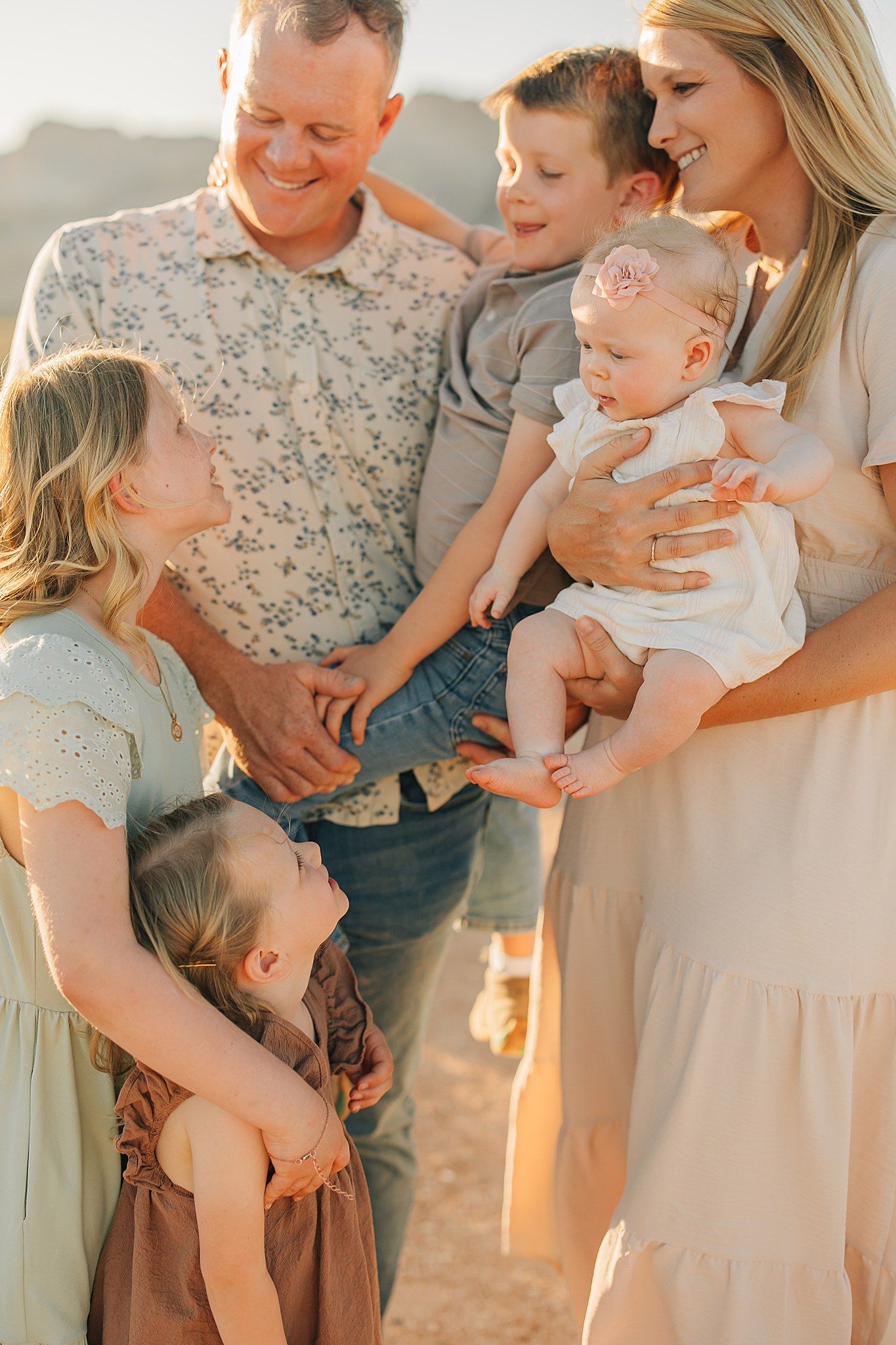 A happy family of five poses outdoors at golden hour, with parents holding children in a warm, candid moment.