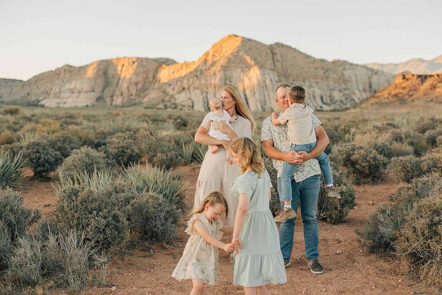 Family posing in desert landscape with red rock mountains at golden hour, parents holding young children in light clothing.