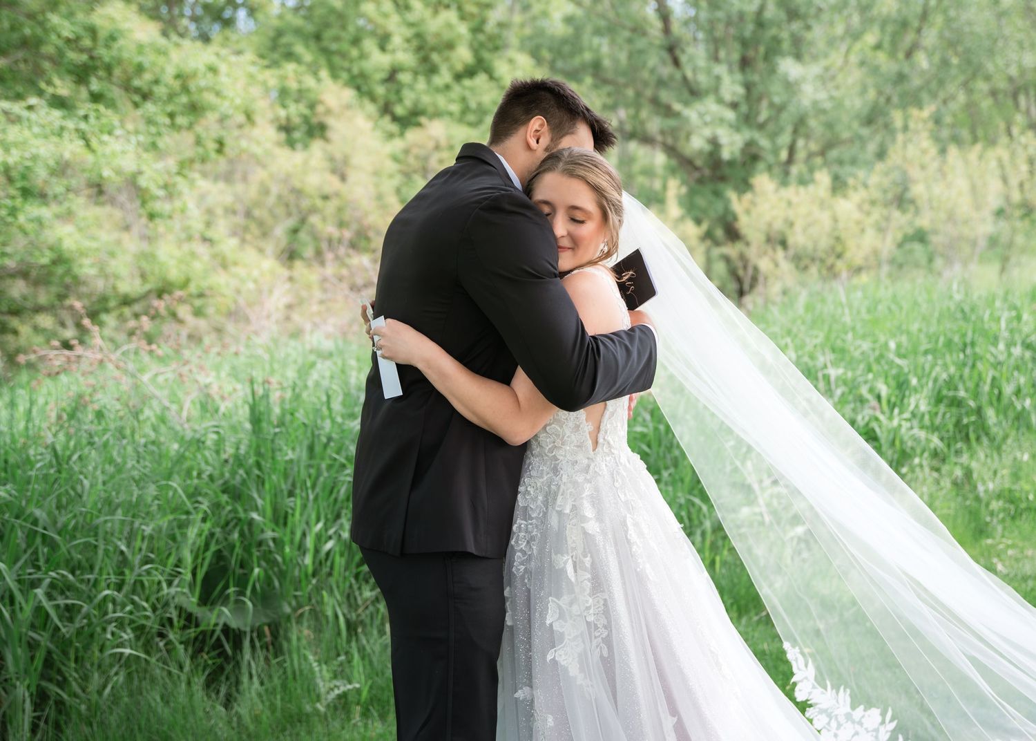 Bride and groom share emotional embrace outdoors, bride in lace gown with veil, surrounded by lush green nature.