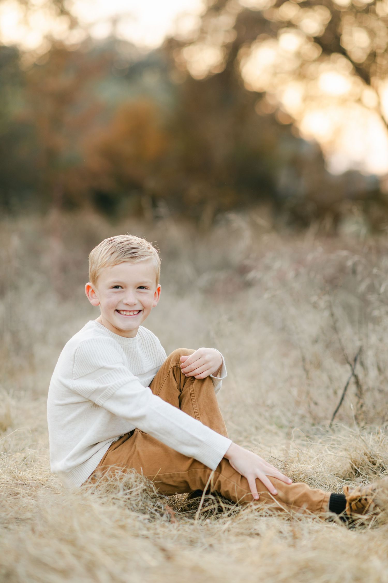 Smiling blonde boy sitting in golden dry grass field wearing white shirt and tan pants during golden hour.