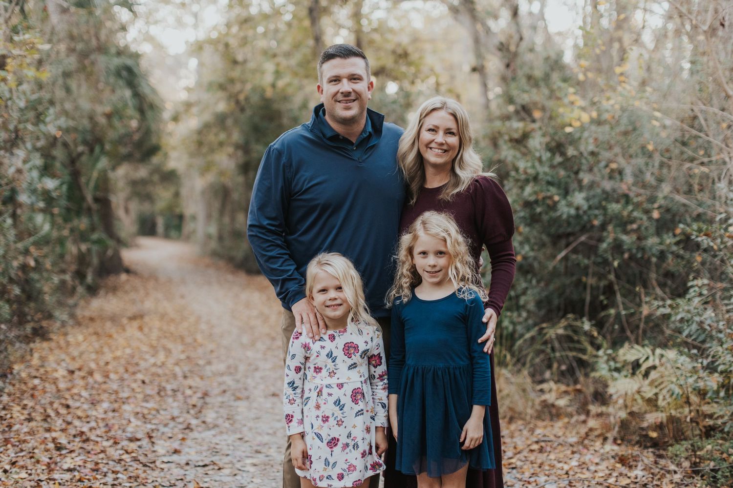 Hilton Head family in family photography session smiling by scenic autumn forest path, parents with two young blonde girls in fall foliage.