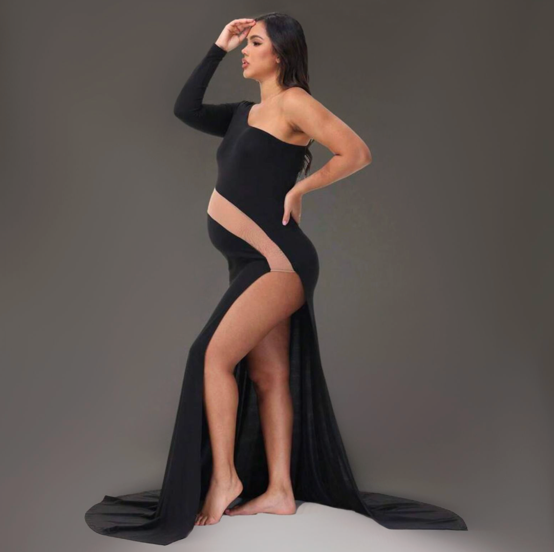 Pregnant woman posing in elegant black asymmetric maternity gown with flowing train and thigh-high slit on gray background.