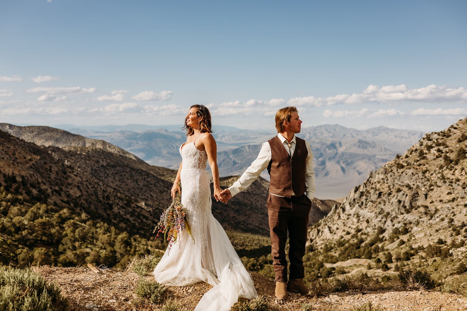 Bride and groom hold hands on a rocky mountain overlook with sweeping valley views on their elopement day.