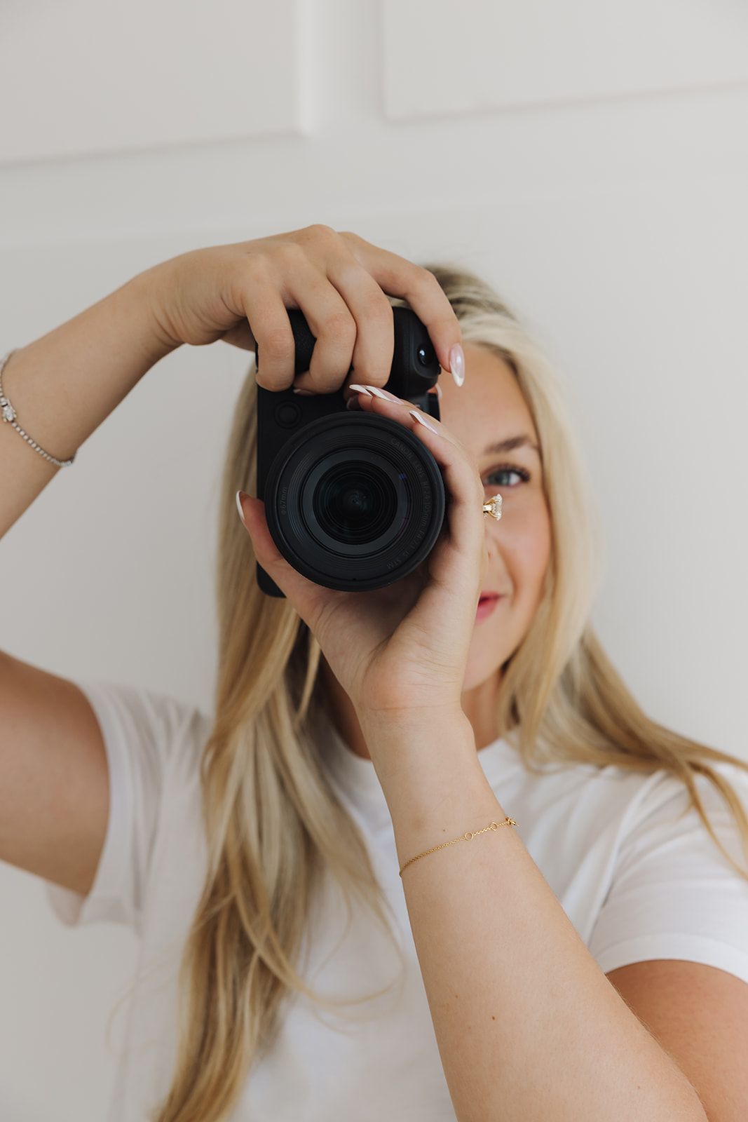 Blonde woman in white t-shirt holding a black DSLR camera up to her eye, wearing gold and silver bracelets.