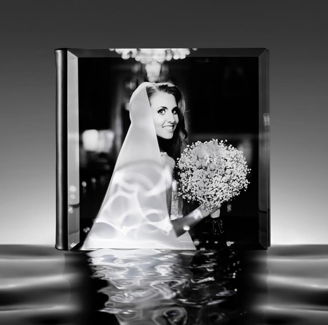 Black and white photo of a smiling bride in veil holding a bouquet, displayed in a reflective glass cube.