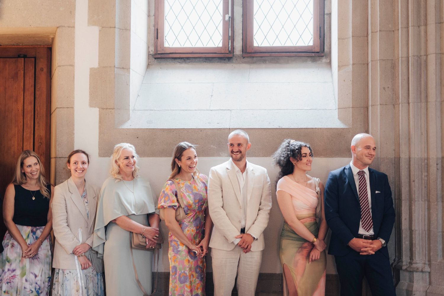 Seven elegantly dressed wedding guests smiling and posing together in a grand stone building.