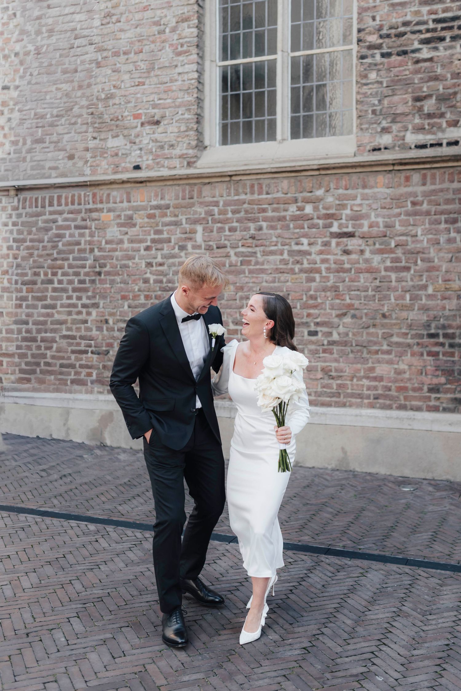 Bride in white midi dress holding bouquet and groom in black tuxedo share a joyful moment on cobblestone street.