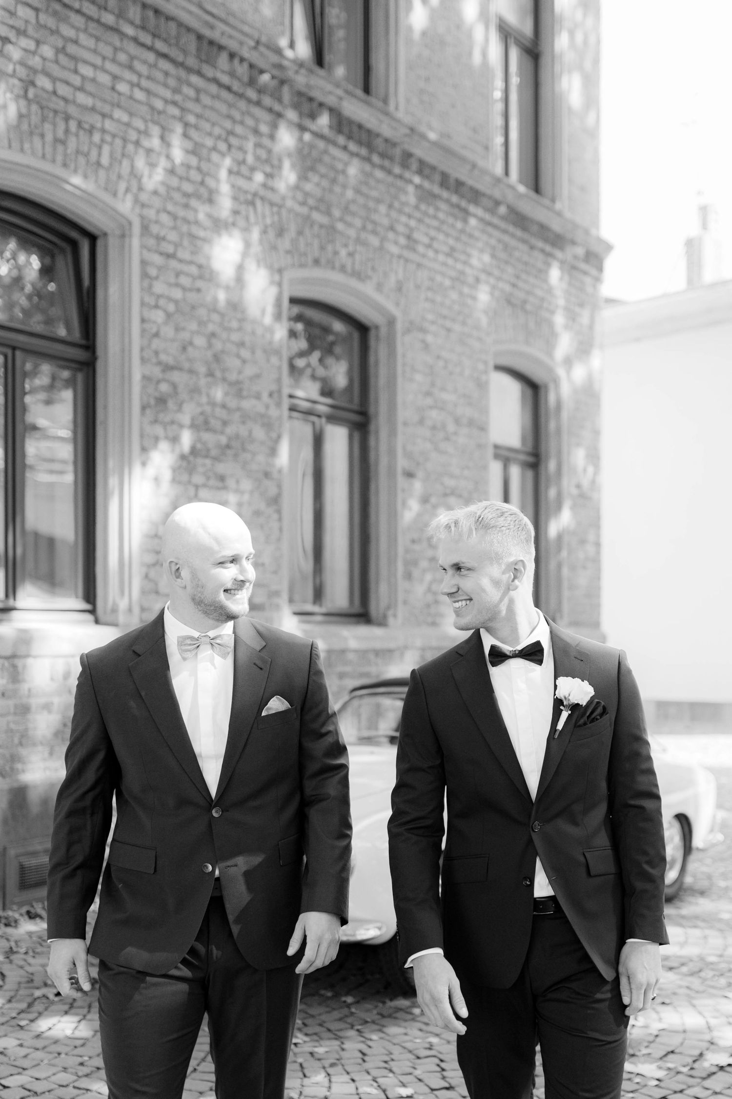 Two grooms in black tuxedos smile at each other outside a brick building on their wedding day.