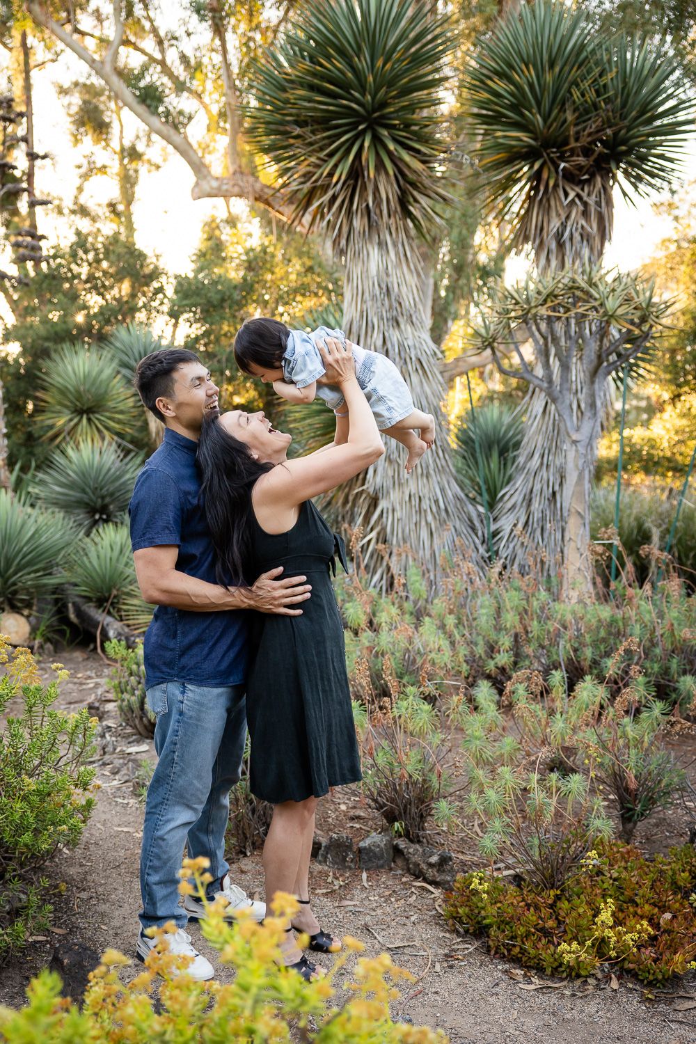 Bay Area family posing in a lush botanical garden, mother lifting baby while father stands beside them, surrounded by desert plants.