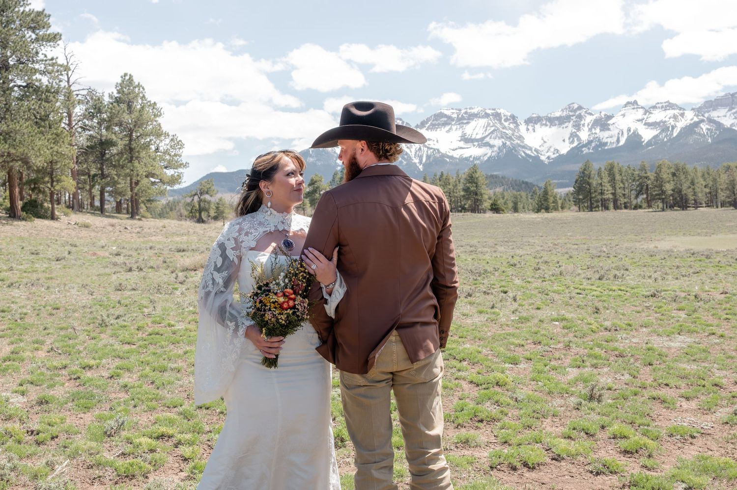 Bride and groom in western attire stand in  meadow, groom wearing cowboy hat, snow-capped peaks of the San Juan Mountains in the background.