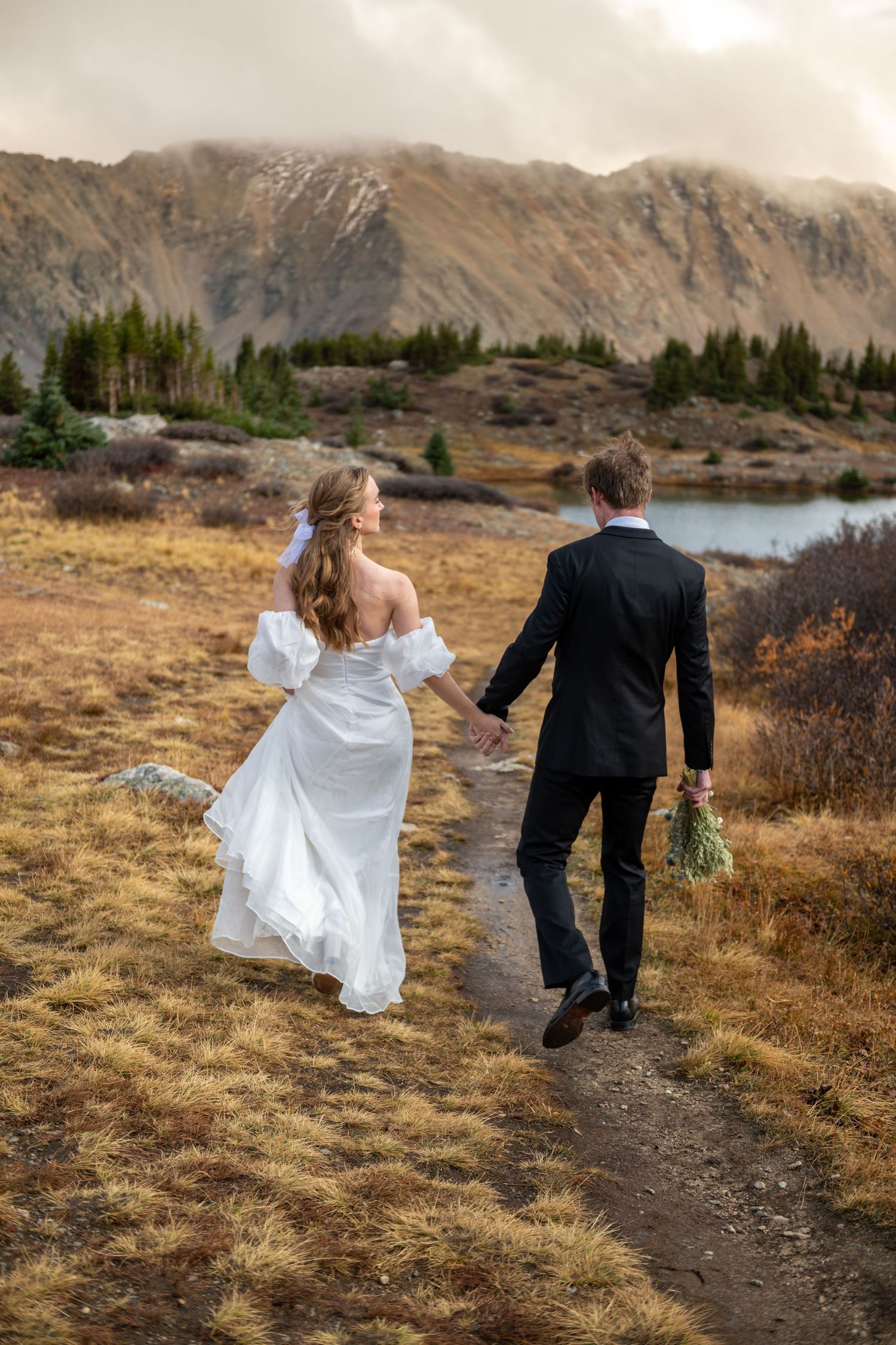 Bride and groom walk hand-in-hand on a mountain trail with a lake and misty peaks in the background.