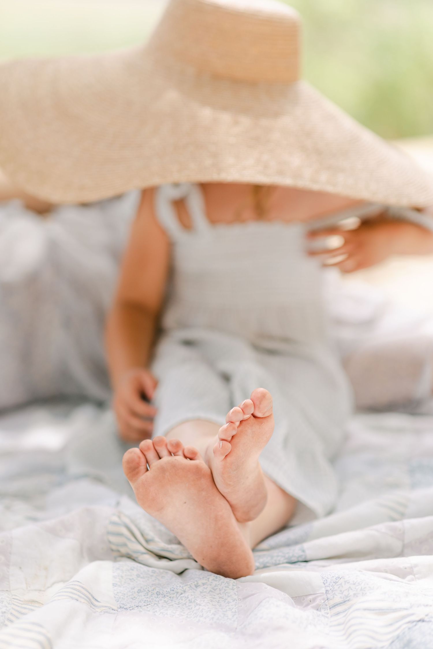 A young girl in a light blue dress and oversized straw hat sits barefoot on a blanket outdoors.