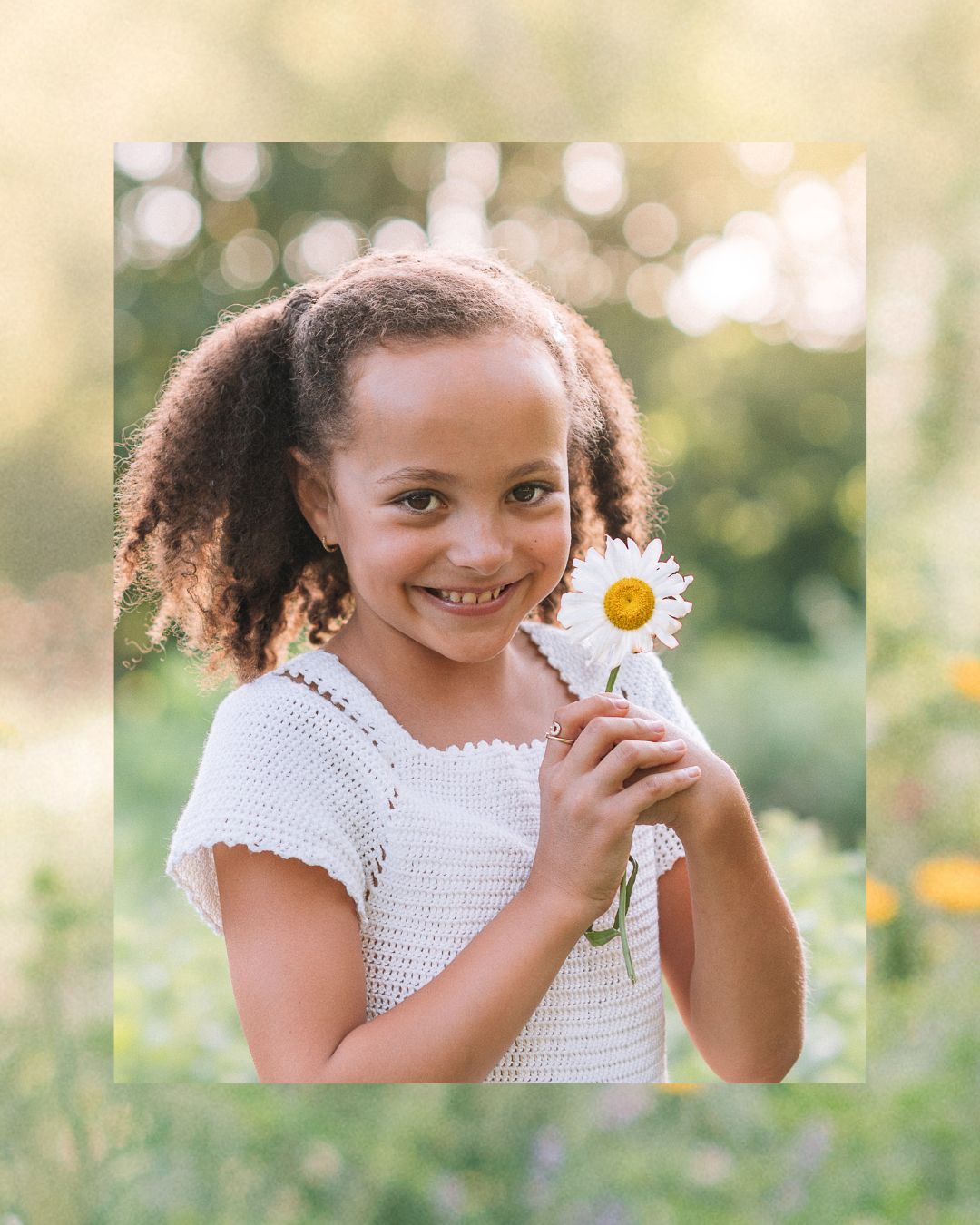 A smiling young girl in a white crochet dress holds a daisy flower outdoors in a sunny green garden.