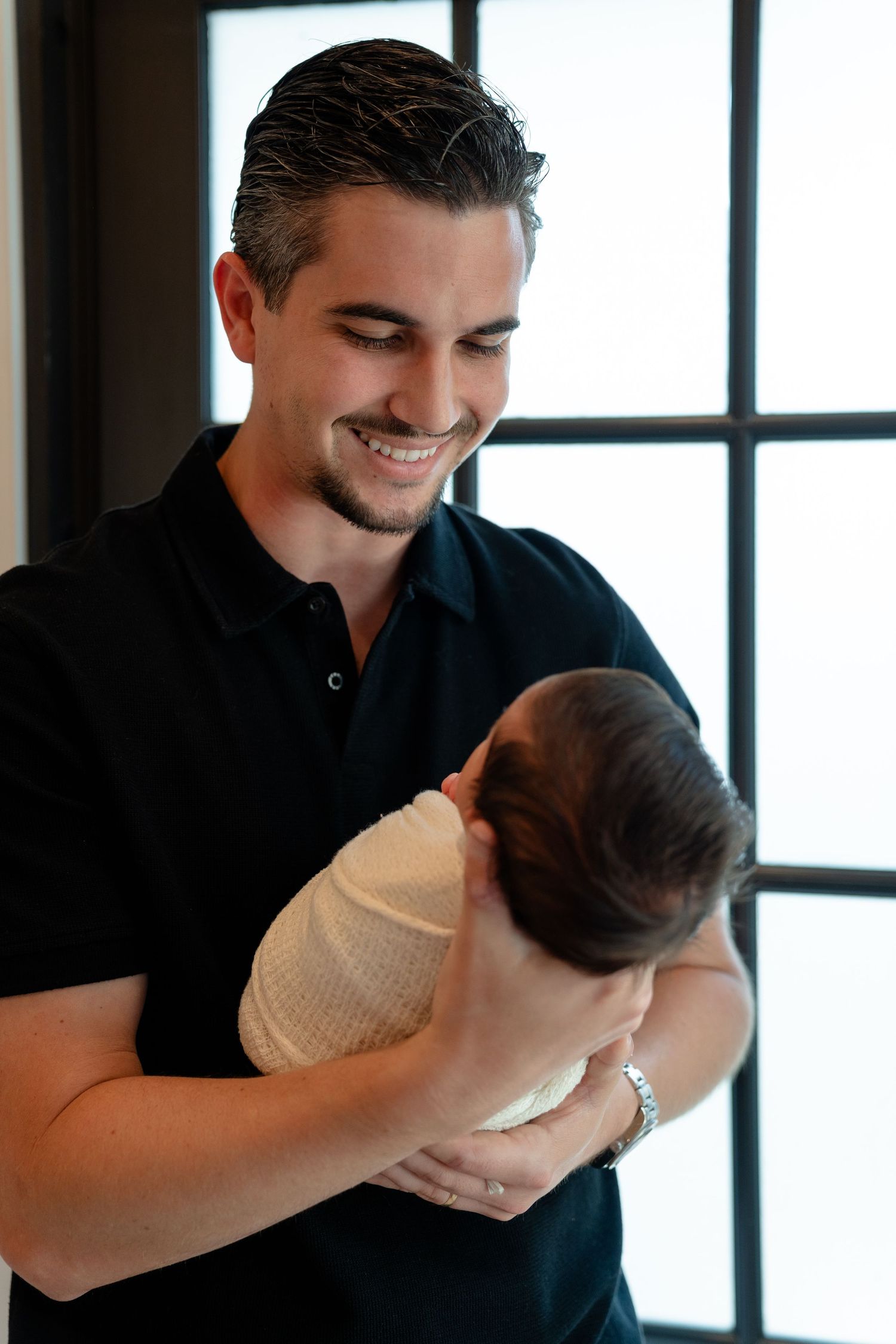 Smiling young man in black polo shirt holding a swaddled newborn baby near a bright window indoors.