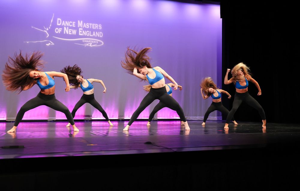 Contemporary dance group performing dynamic hair-flipping choreography on purple-lit stage.