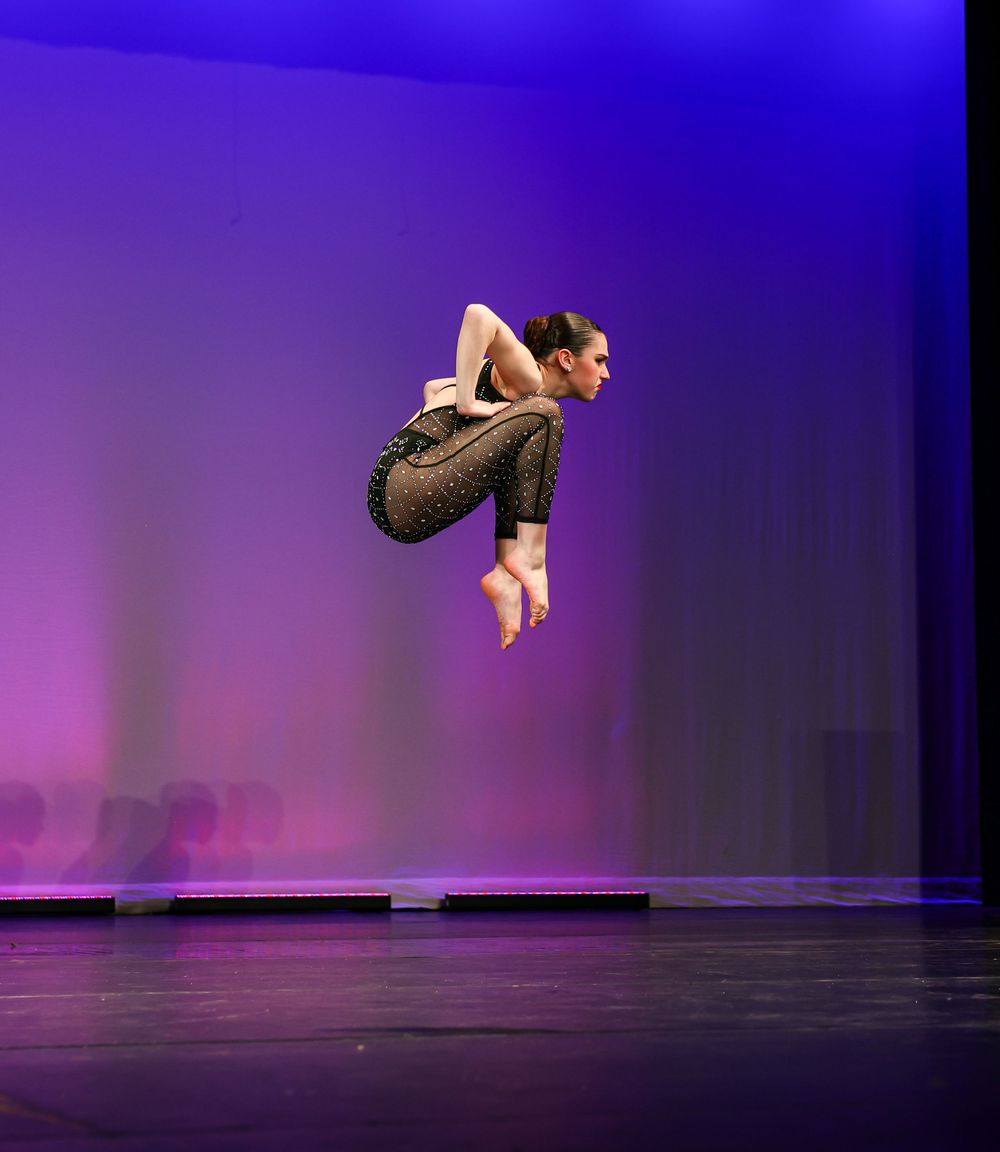 A dancer performs a dynamic leap against a purple-lit stage backdrop during a contemporary dance performance.
