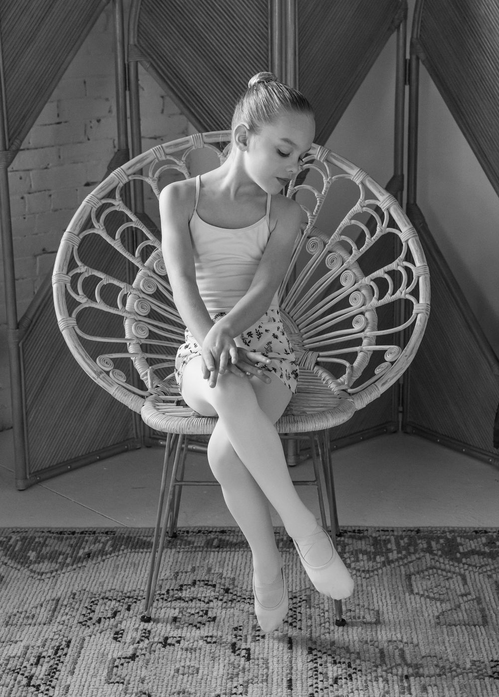 A young ballet dancer in white leotard sits gracefully in a vintage peacock chair on a patterned rug in black and white.