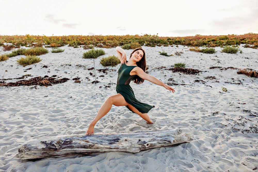 Dancer in green dress performs graceful pose on driftwood at beach during sunset with coastal vegetation in background.