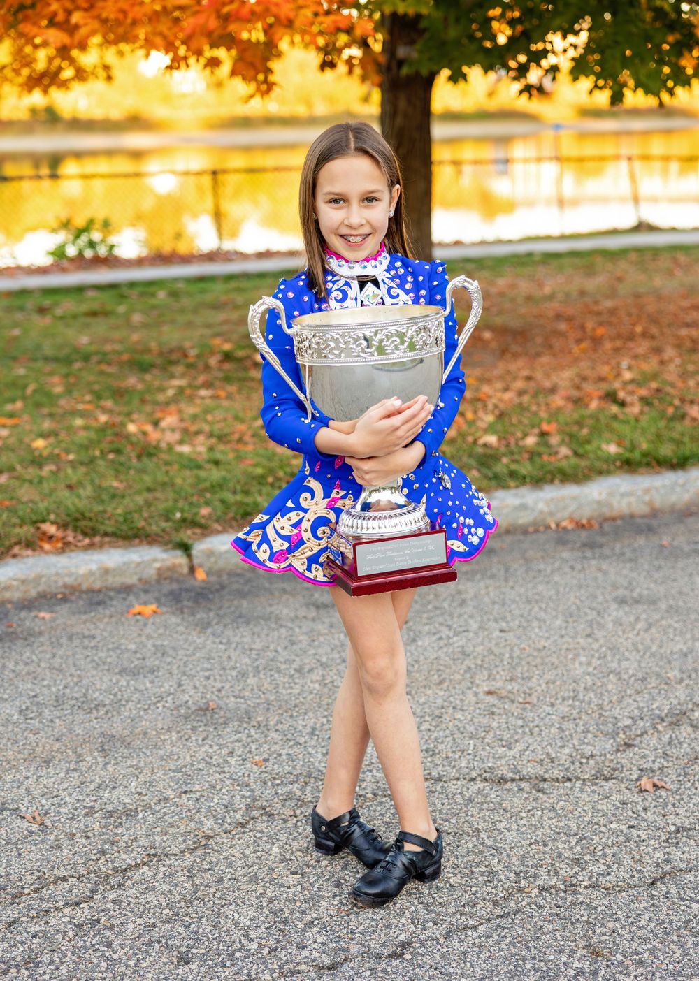 Irish dancer holding trophy while wearing blue competition dress with autumn scenery behind.