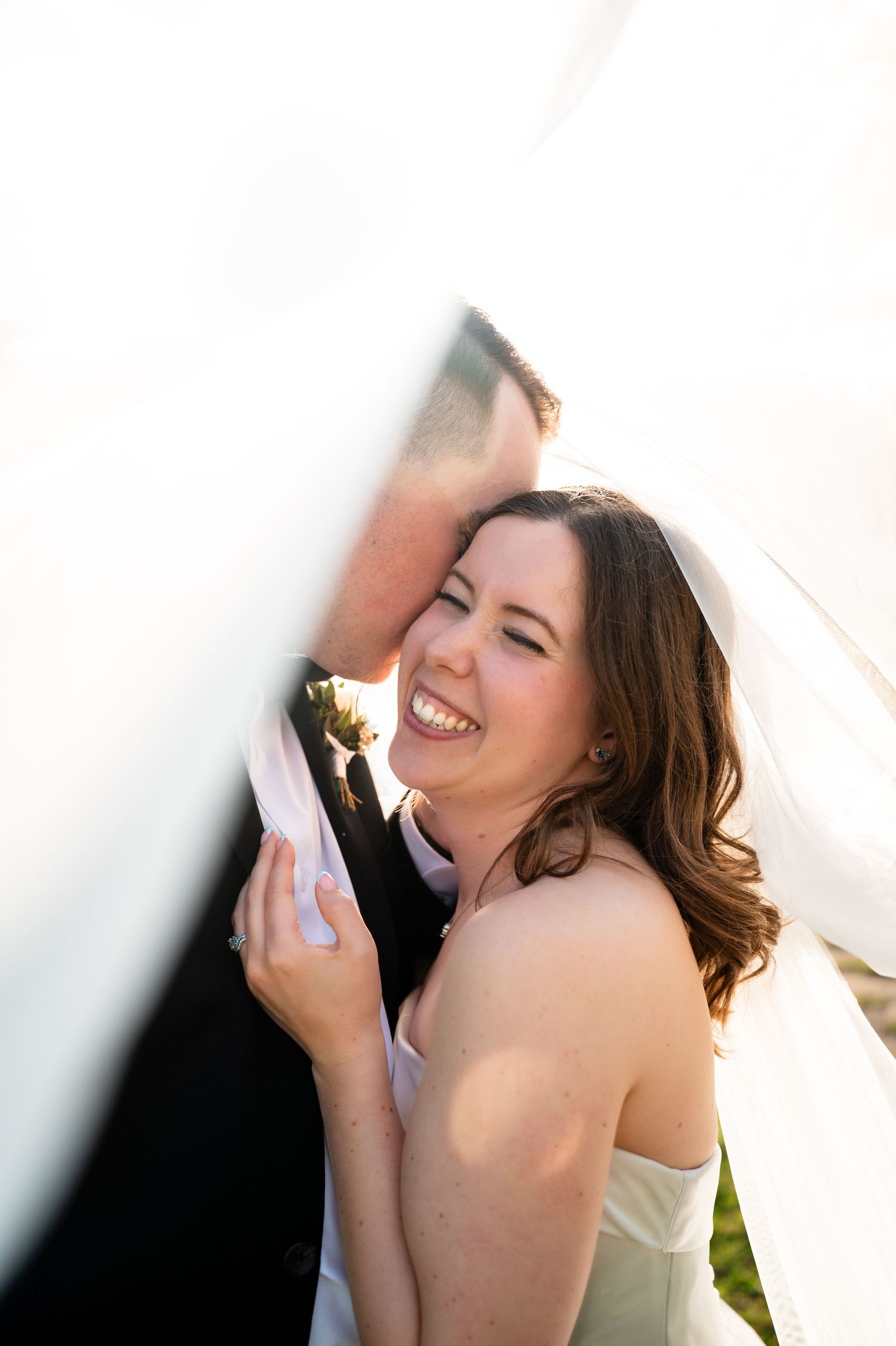 Bride laughing joyfully as groom kisses her cheek, framed by flowing white wedding veil in romantic outdoor portrait.
