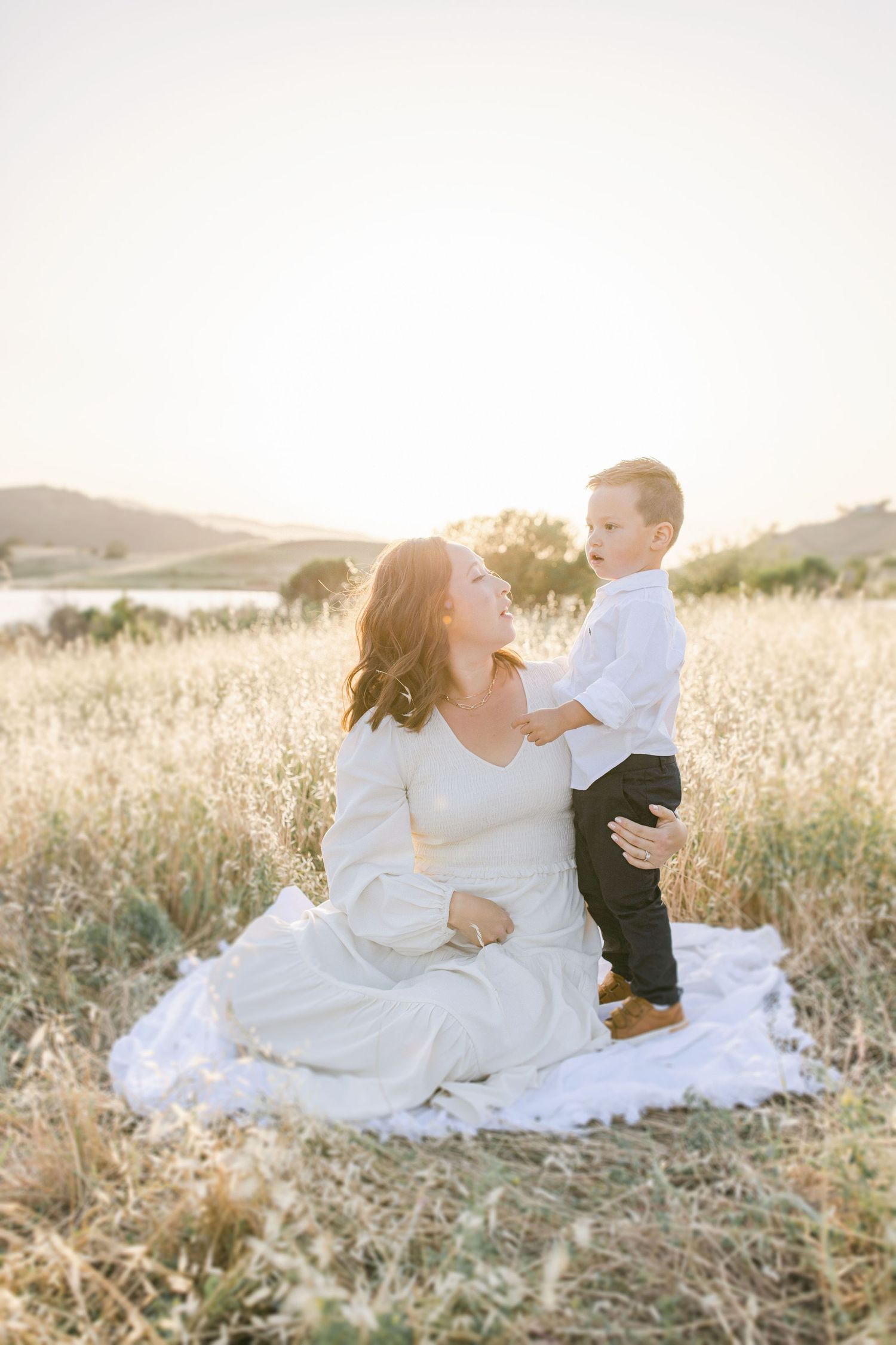 A mother in a white dress sits on a blanket in a golden field, smiling up at her young son during sunset.