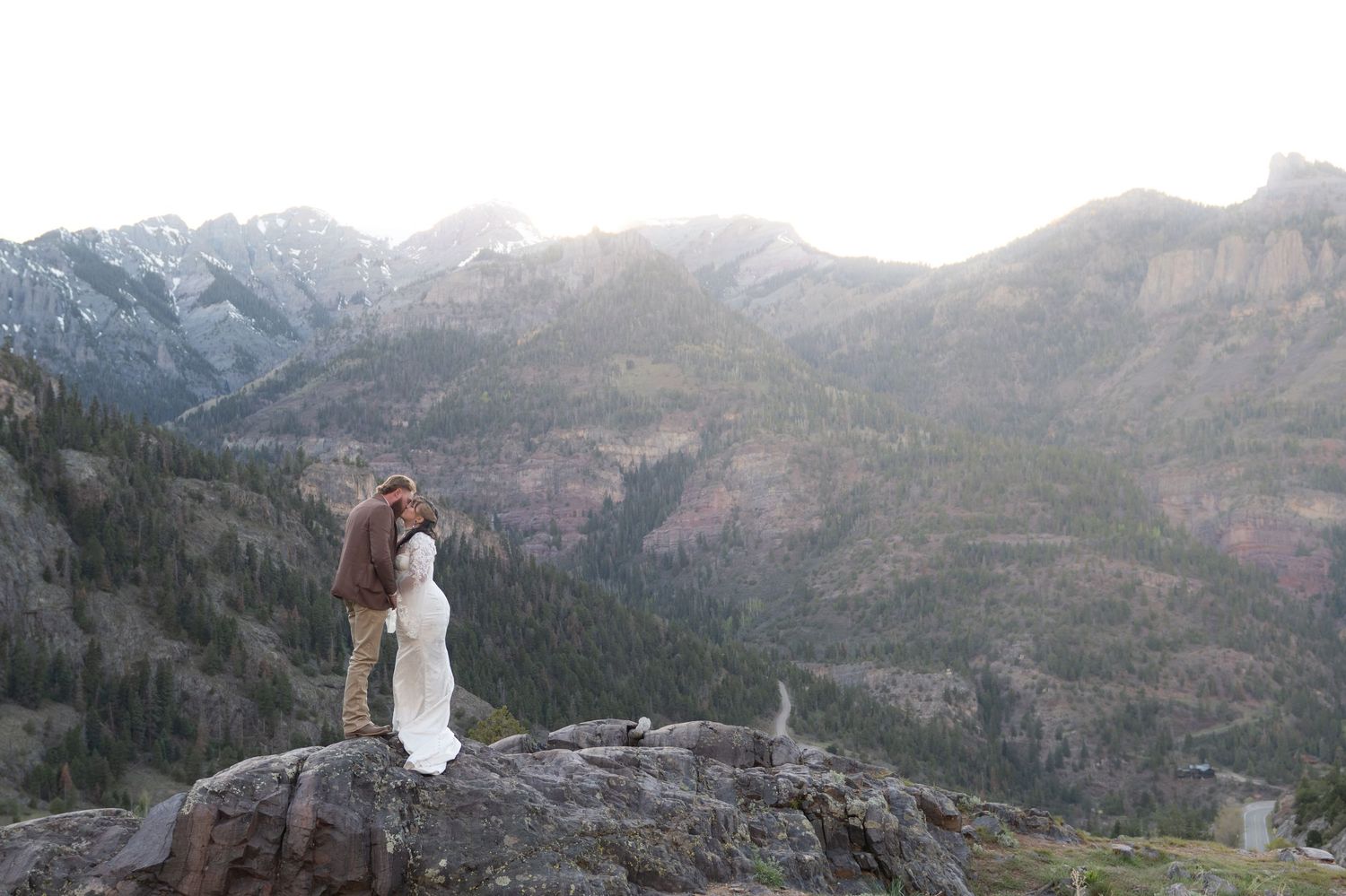 Couple sharing a romantic kiss on a rocky mountain summit with stunning valley and peaks backdrop at sunset.
