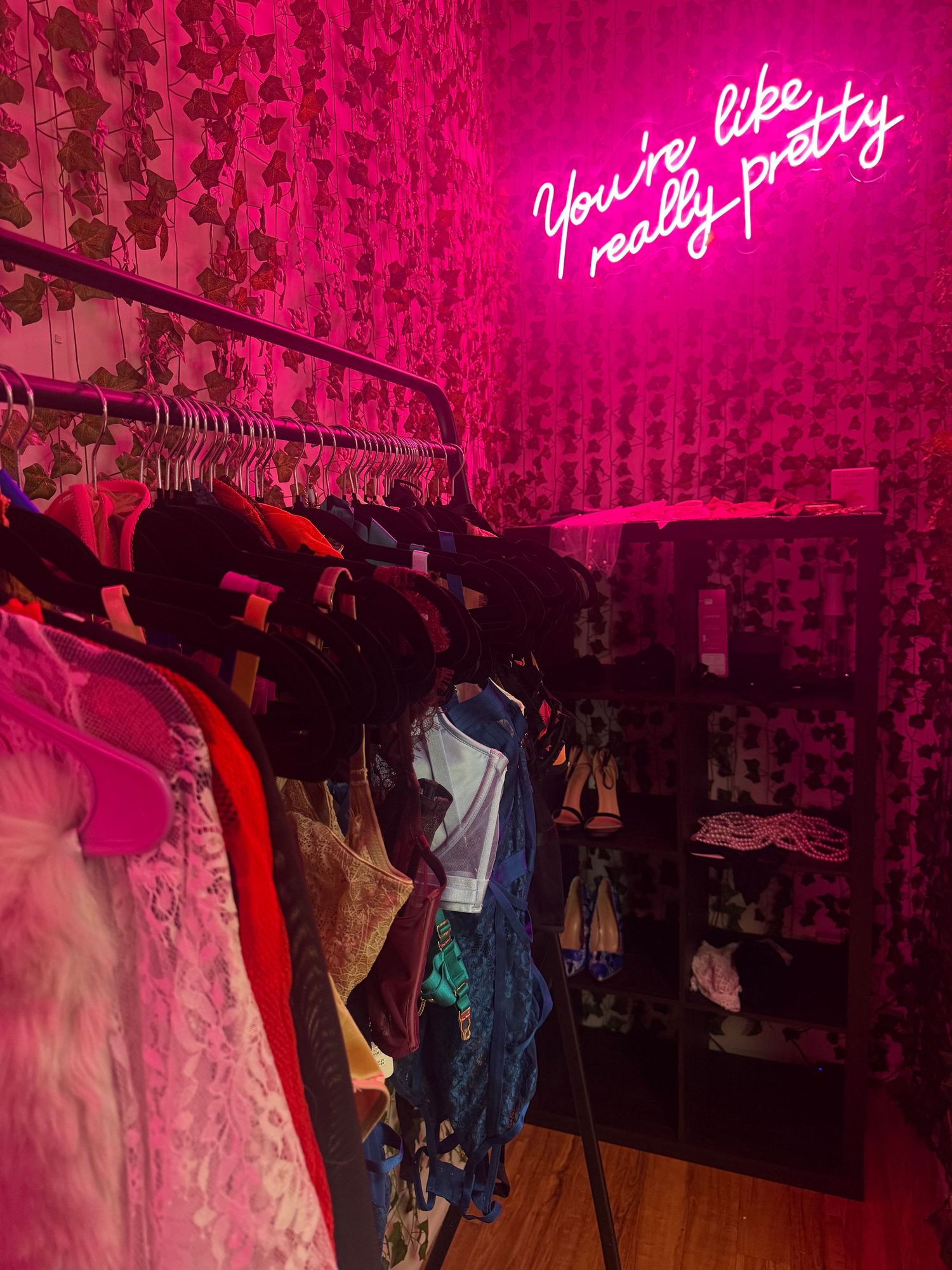 Pink neon sign reading you're like really pretty above clothing rack in boutique with floral wallpaper.