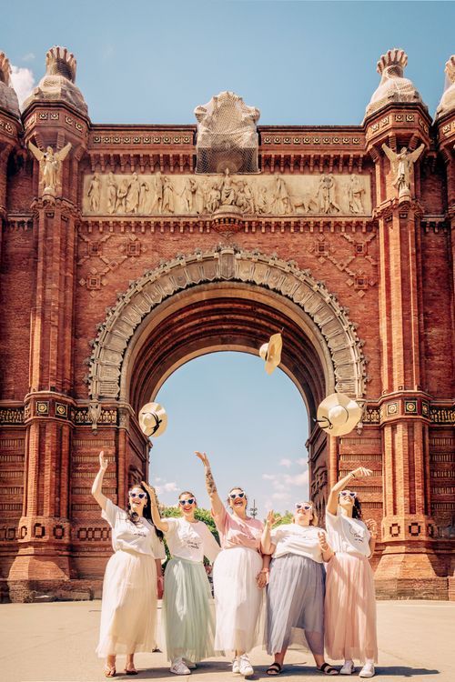 Five women in matching outfits tossing straw hats in front of the Arc de Triomf in Barcelona, Spain.