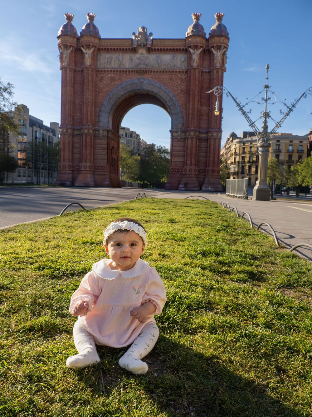 Baby girl in pink outfit sitting on grass in front of Arc de Triomf, Barcelona, on a sunny day.