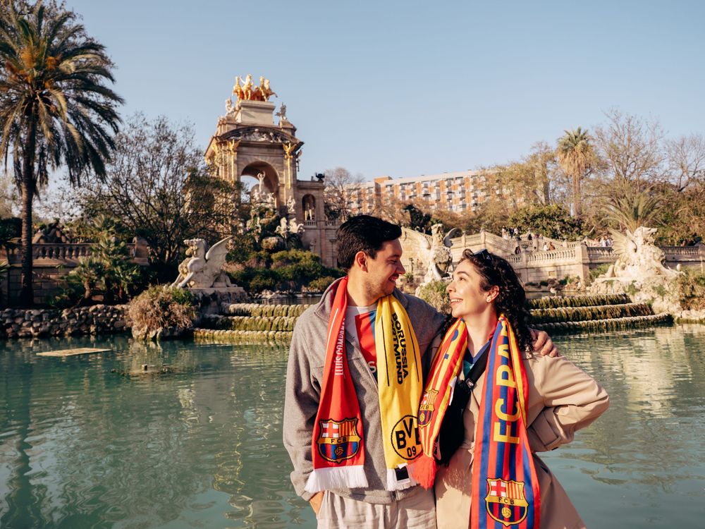 Couple wearing FC Barcelona and BVB scarves smiling at each other by a fountain in Parc de la Ciutadella.
