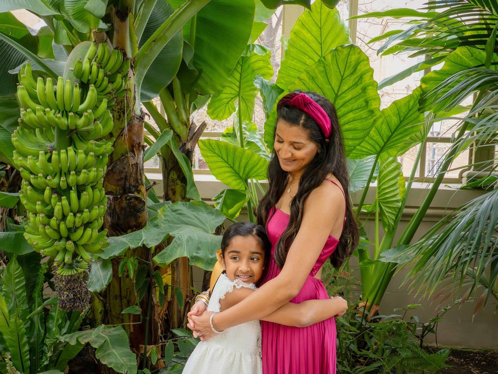 A woman in a pink dress hugs a young girl in white amid lush tropical banana and elephant ear plants.