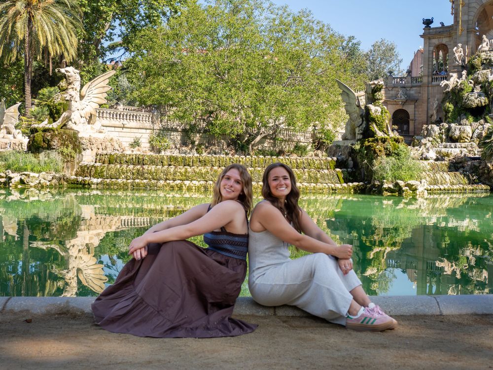 Two women pose back-to-back by a ornate fountain pool in a lush garden, surrounded by sculptures and greenery.