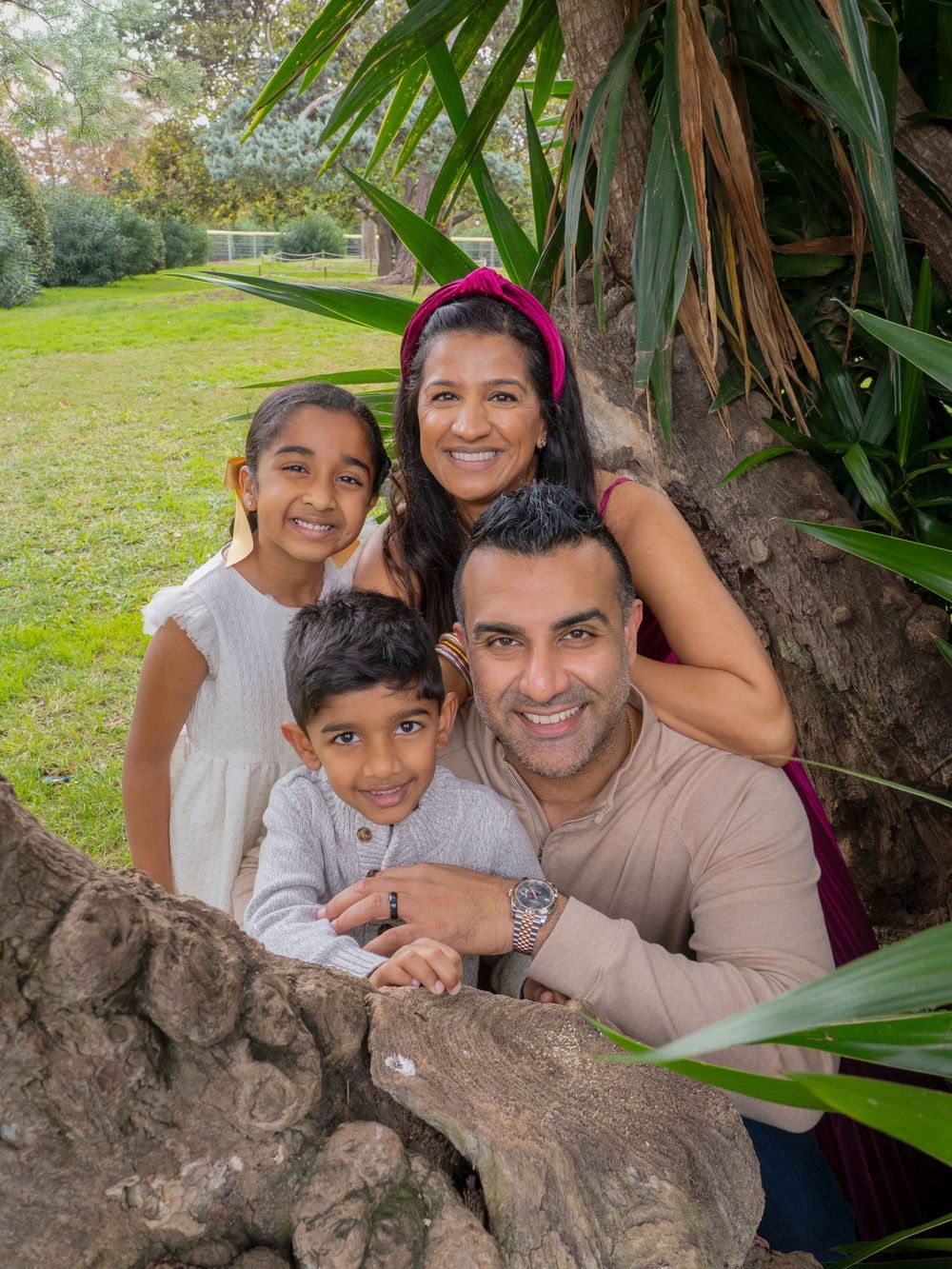 A smiling South Asian family of four poses together by a large tree in a lush green park setting.
